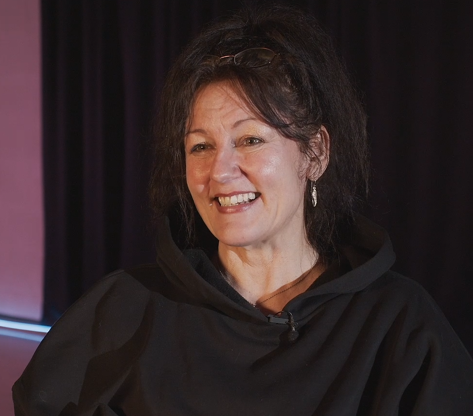A person smiling and wearing a black hoodie, sitting in front of a dark background.